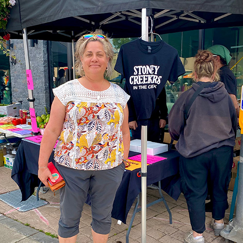 Juanita enjoying the market place on the street during apple Fest in front of a shirt that says Stoney Creekrs